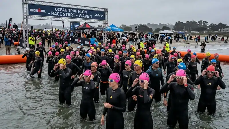 Triathletes in wetsuits standing in shallow water wearing tinted goggles under an overcast sky
