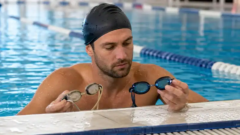 Competitive swimmer comparing old and new swim goggles at a pool wall