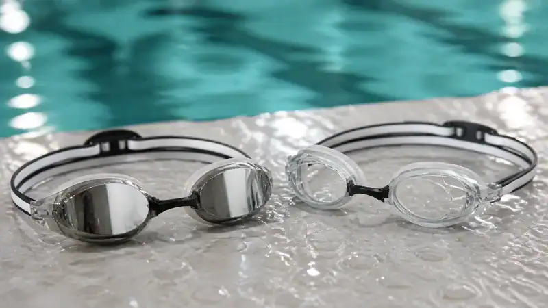 Mirrored and clear racing goggle lenses rest side by side at the edge of an indoor pool.